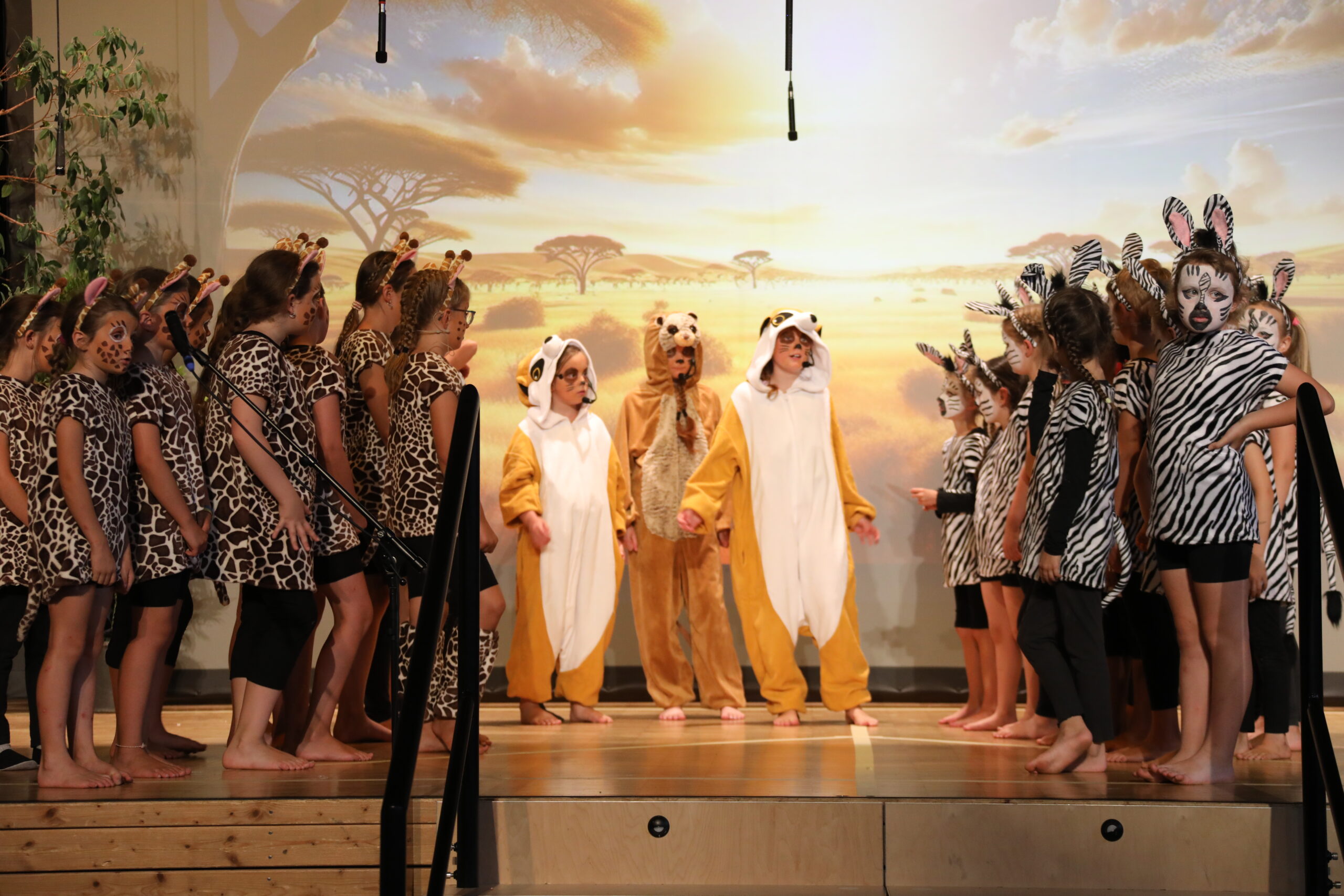 Kinder stehen in Tierkostümen auf der Bühne und singen. Als Zebra, Giraffe usw. Im Hintergrund sieht man die Savanne.