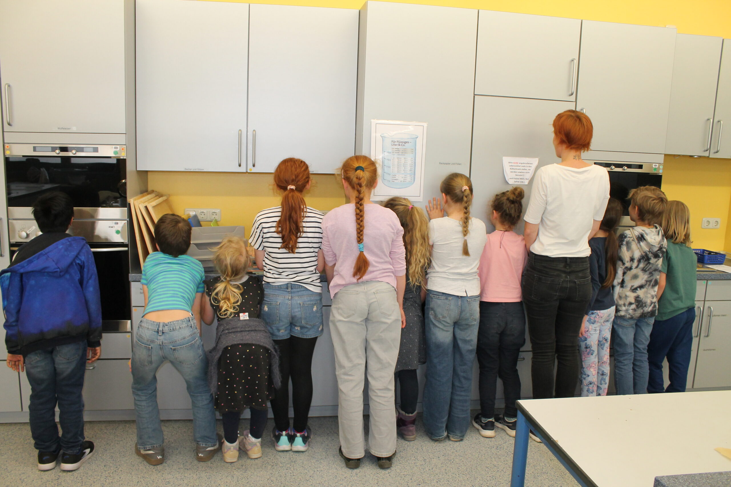 11 Kinder und eine Lehrerin stehn vor einer Küchenzeile, man sieht sie von hinten.