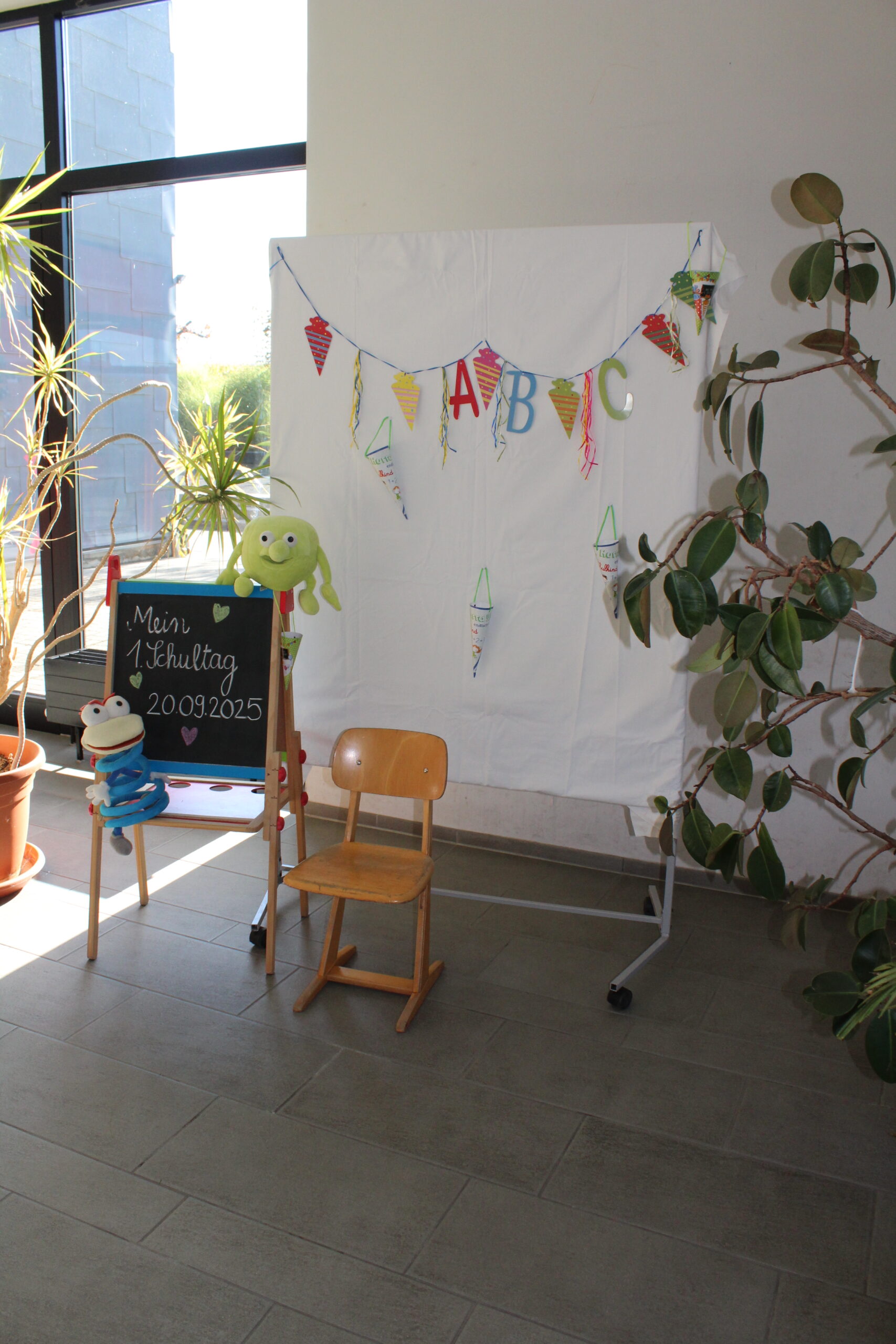 Auf einer Kinderspieltafel steht: Mein 1. Schultag 20.09.2025. Daneben steht ein Stuhl, im Hintergrund hängt eine ABC Girlande.