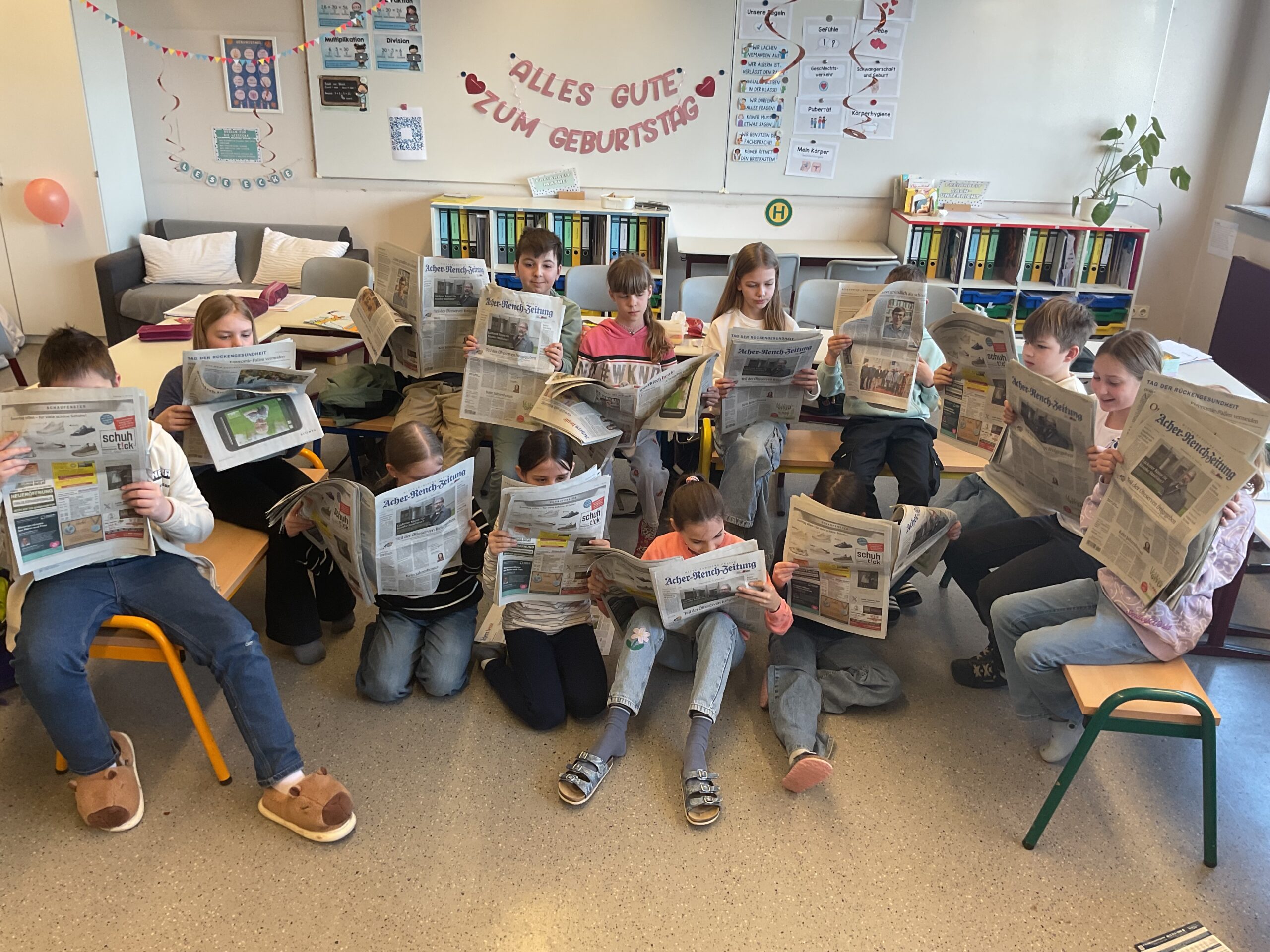 14 Kinder sitzen im Klassenzimmer und lesen Zeitung.
