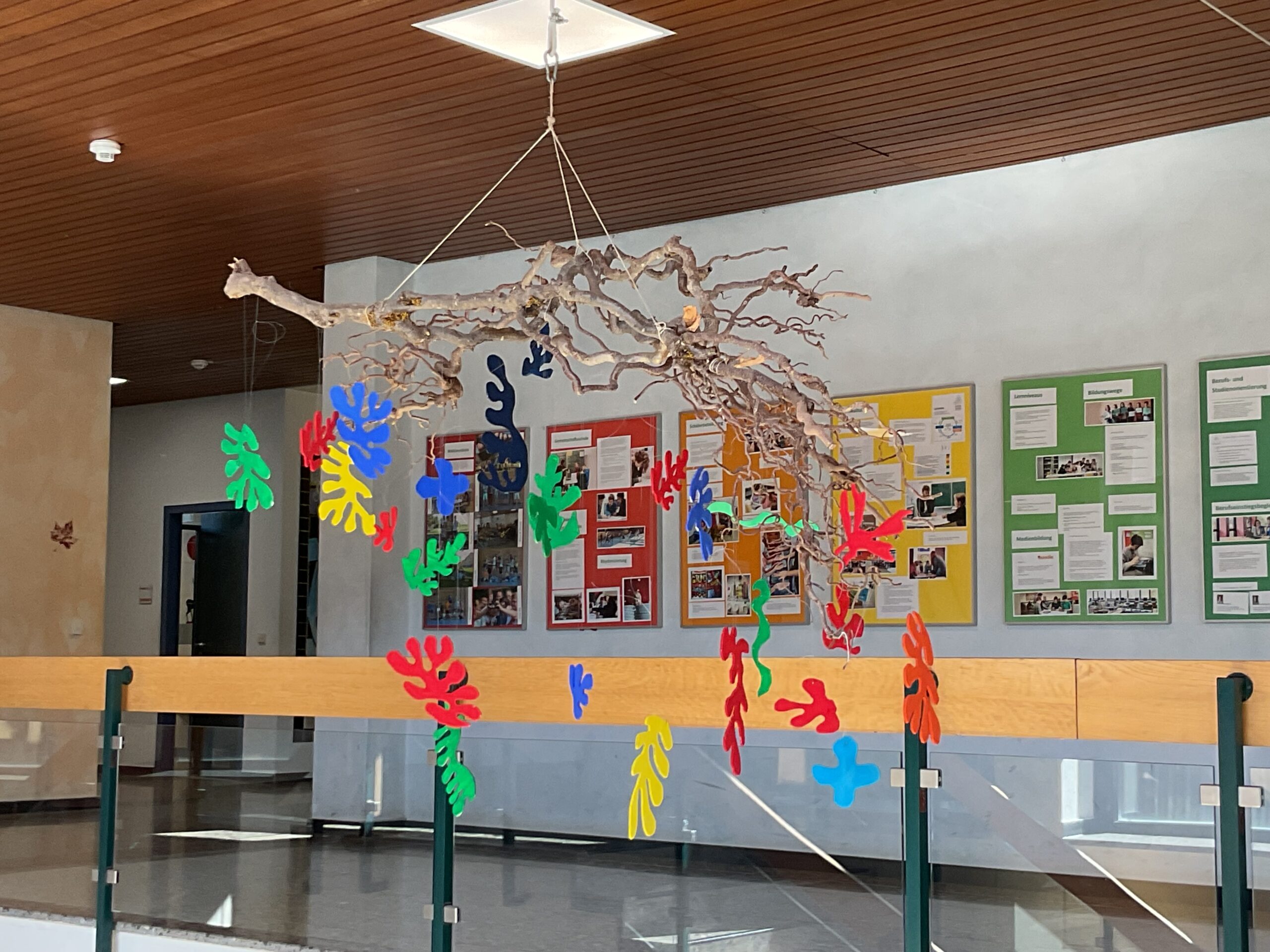 Man sieht einen Zweig im Schultreppenhaus aufgehängt. An dem hängen aus buntem Papier ausgeschnittenen Blätter.