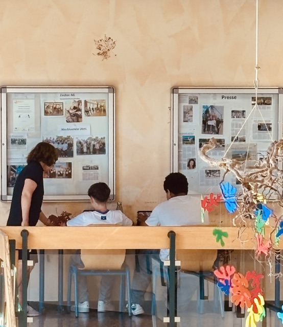 Ein Lehrer steht neben zwei Schülern die an Schreibtischen im Schulflur sitzen und erklärt etwas.