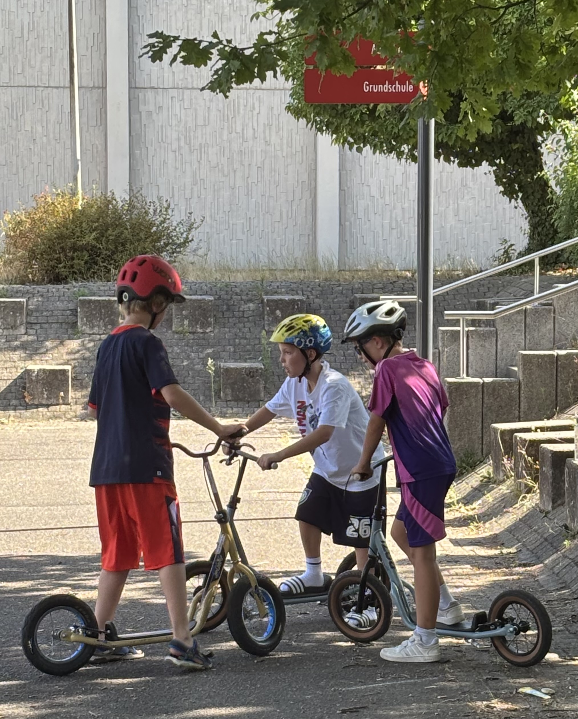 3 Kinder stehen mit Ihren Rollen auf dem Schulhof zusammen und unterhalten sich.