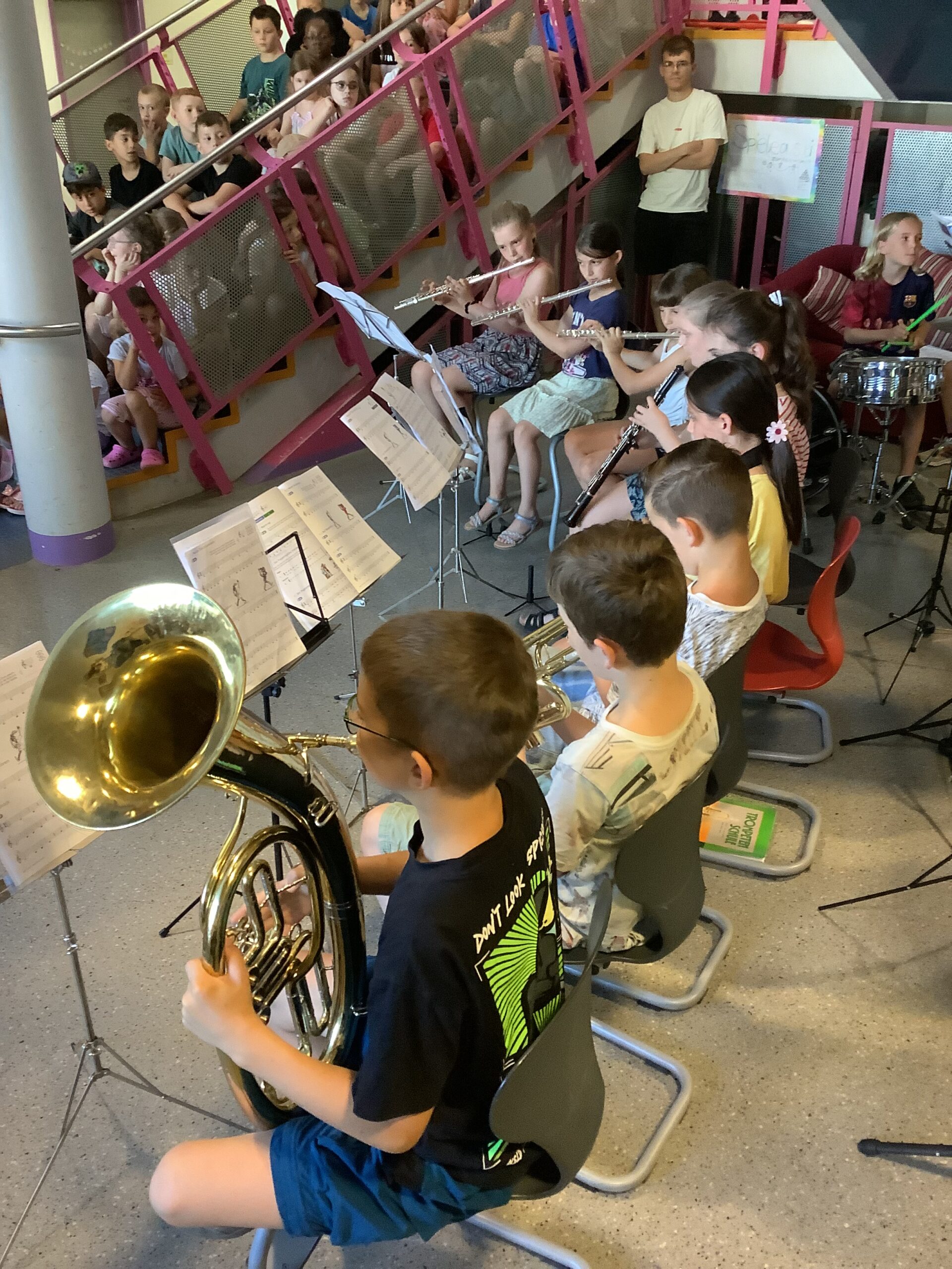 Ein Kinderblasmusikorchester spielt etwas vor. Kinder sitzen auf der Treppe und hören zu.