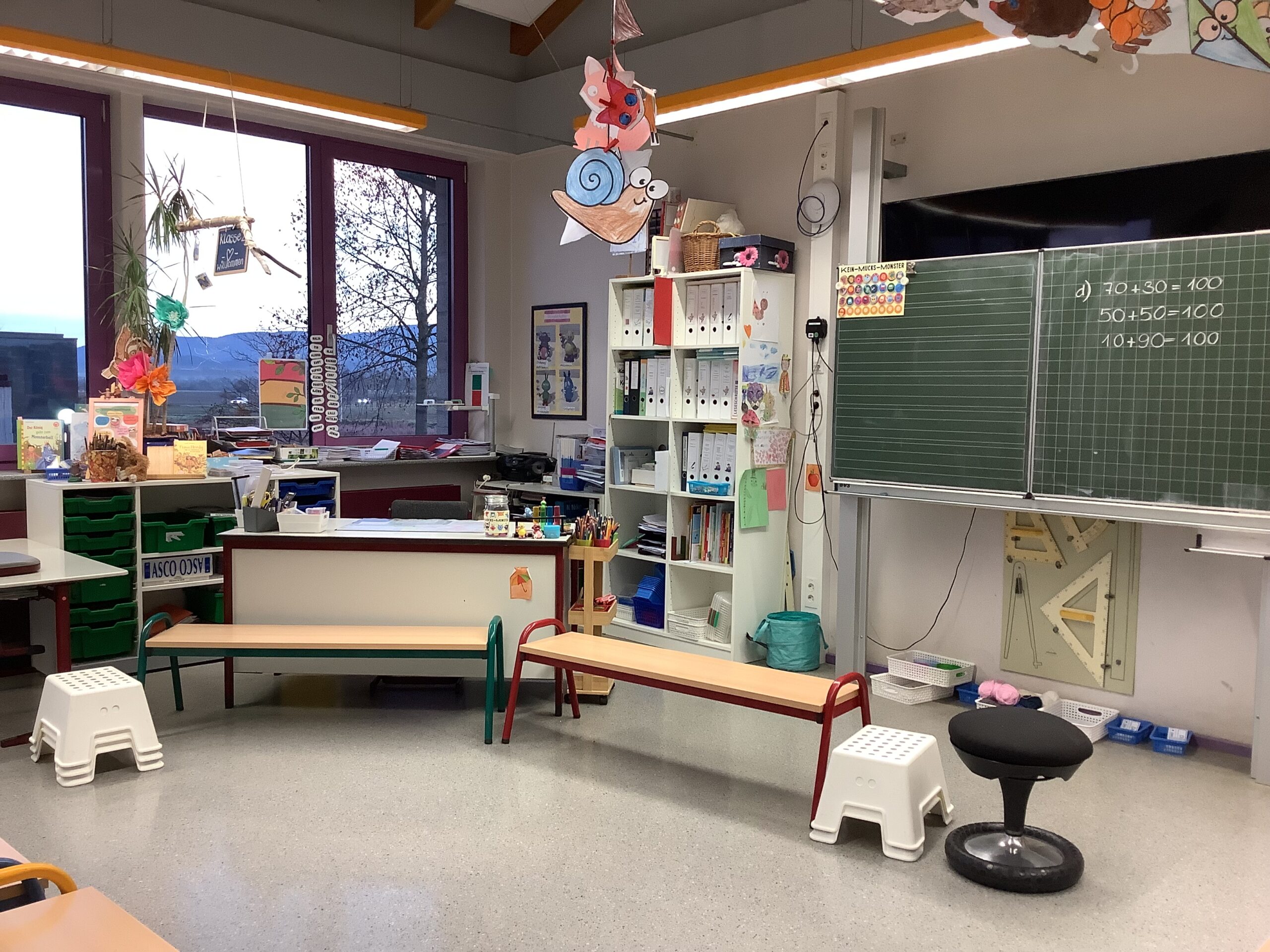 Klassenzimmer mit Lehrerpult, Bänken und Hocker für einen Stuhlkreis.